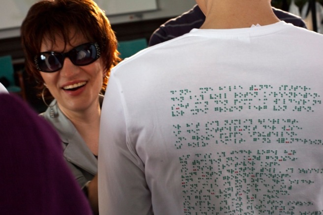 Links lacht uns eine Frau mit Sonnenbrille an. Ein sehr sympathisches, strahlendes Lachen. Rechts neben ihr ist der Rücken eines Mannes mit weißem T-Shirt, auf dem ein Text in grüner Punktschrift mit vereinzelten roten Punkten steht. (Karl Ahl)