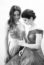 Zwei junge Frauen in Abendkleidern schauen einander an und befühlen ihre Kleider. Schwarz-Weiß